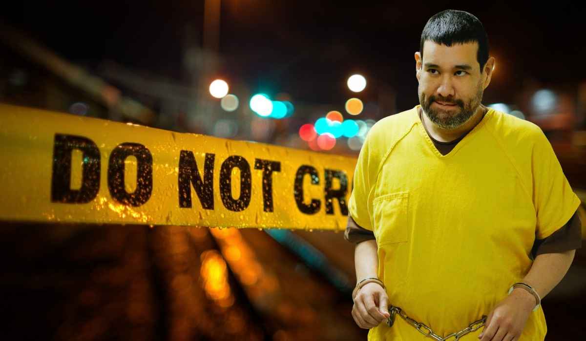 A man in a yellow prison jumpsuit and handcuffs stands in front of blurred crime scene tape reading “DO NOT CROSS