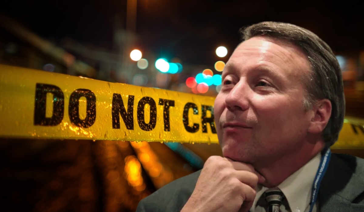 A man in a suit poses thoughtfully in front of blurred crime scene tape reading “DO NOT CROSS.”