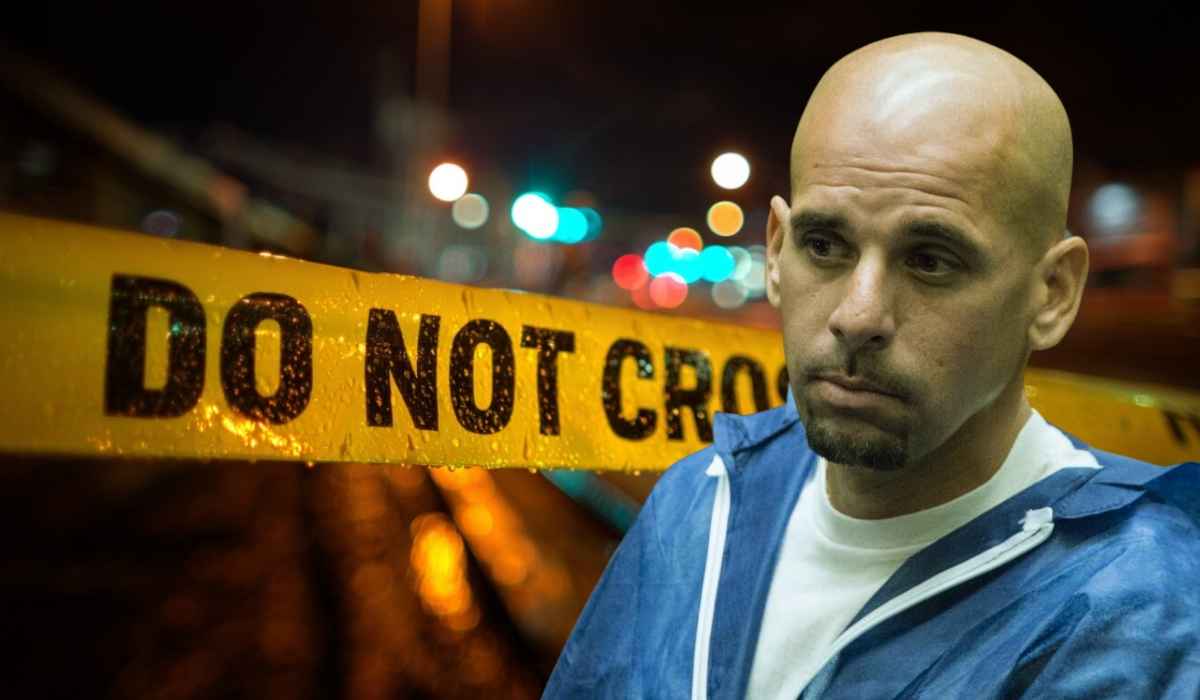A man in a blue jumpsuit stands in front of yellow police tape with “DO NOT CROSS” at a nighttime crime scene